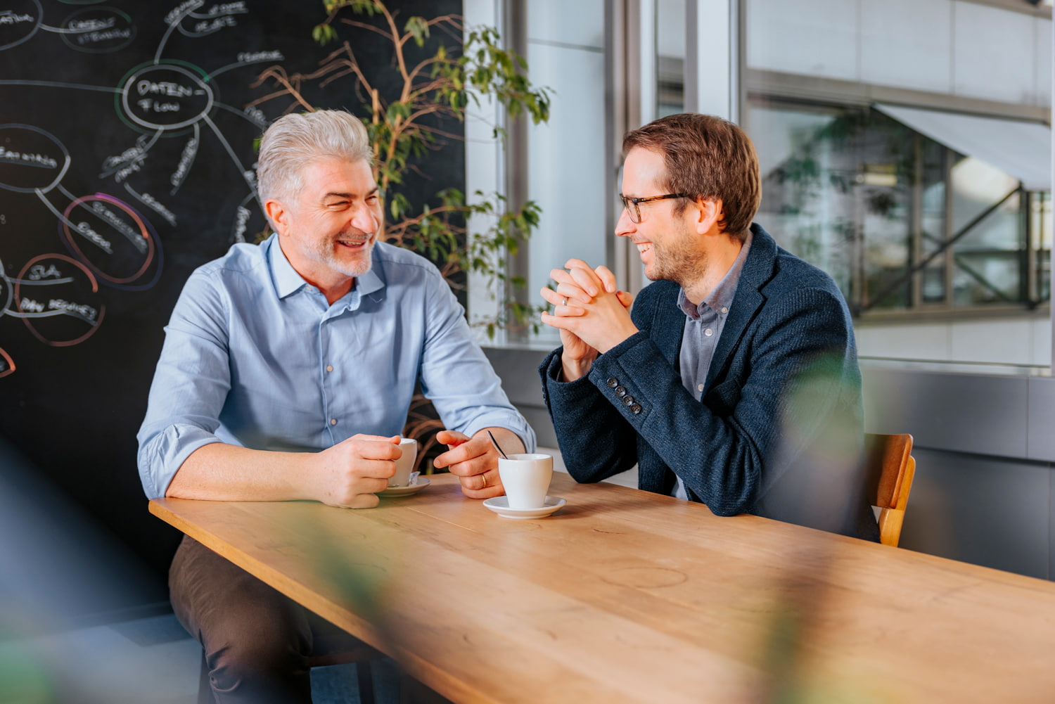 Zwei Männer sitzen an einem Tisch mit einem Kaffee und lachen.