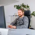 Mann in grauem Pullover sitzt vor einem Bildschirm am Pult