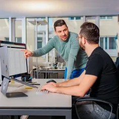 Ein Mann mit schwarzem T-Shirt sitzt am Pult vor dem Laptop und ein anderer Mann erklärt ihm etwas.