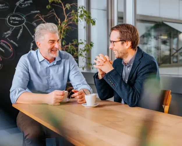 Zwei Männer sitzen an einem Tisch mit einem Kaffee und lachen.