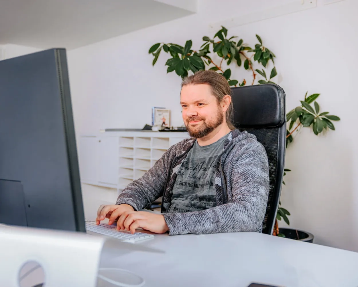 Mann in grauem Pullover sitzt vor einem Bildschirm am Pult