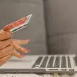 Die Abbildung zeigt eine Nahaufnahme einer Person, die mit einer Kreditkarte in der Hand vor einem Laptop in einem Onlineshop stöbert.