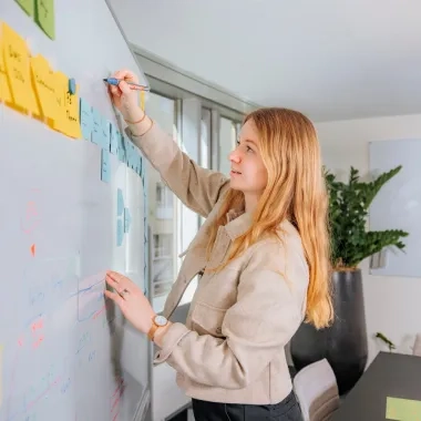 Frau mit blonden Haaren steht vor einem White Board.